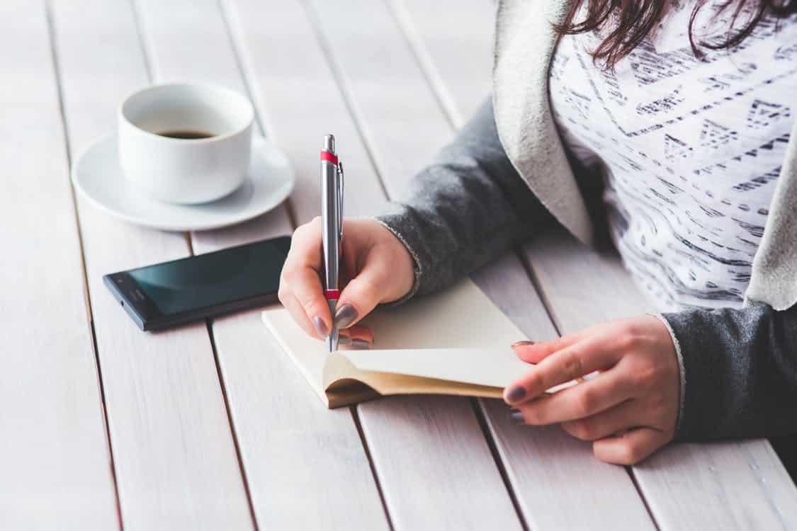אישה כותבת במחברת על שולחן מעץ שיש עליו קפה וסמארטפון