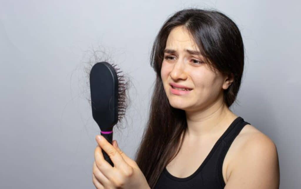 אישה מסתכלת בפנים עצובות על המברשת שיער שלה שמלאה בשערות שנשרו מהראש